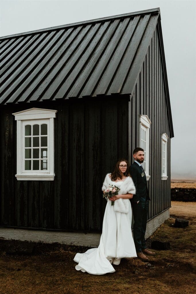 Couple standing outside the Búðir Black Church during an Iceland elopement, with the dramatic black chapel and moody landscape behind them.
