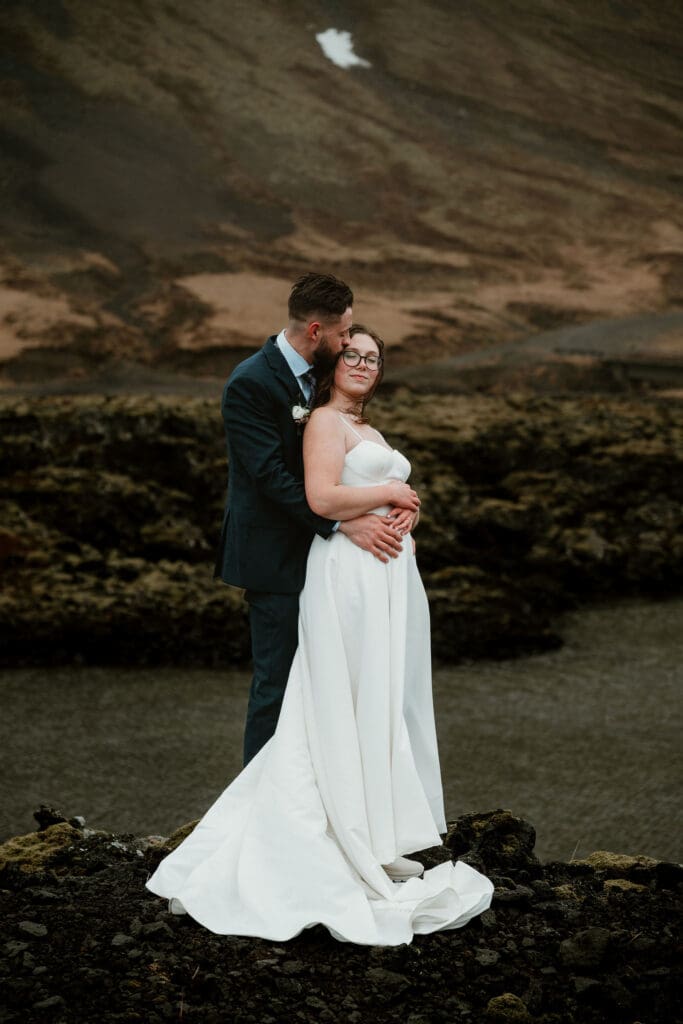 In Snæfellsnes peninsula, a groom hugs his wife from behind, kissing her temple as she closes her eyes, they are surrounded by black lava and mossy landscapes