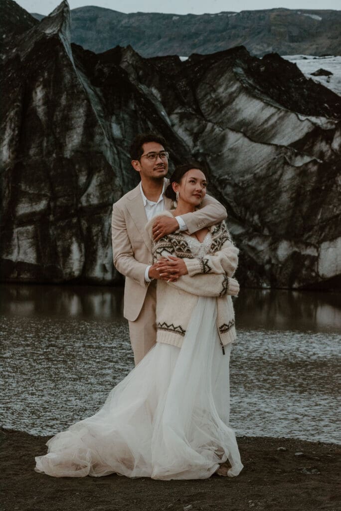 Elopement couple holding each other, facing the camera and looking into the distance in soft, subdued light and an ash-striped glacier behind them