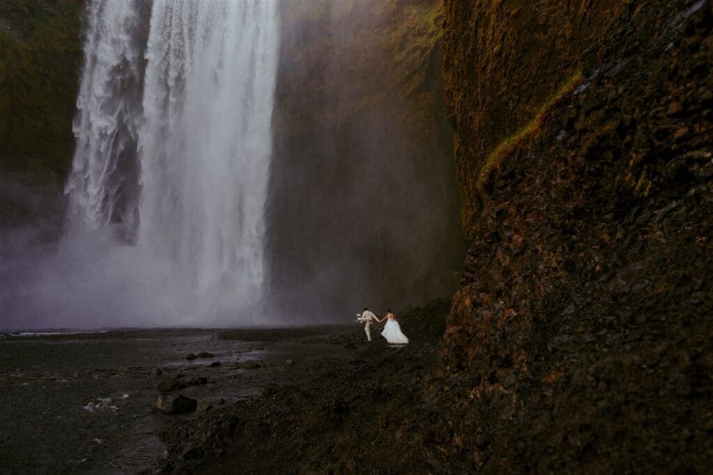 Couple walking hand in hand beside Skógafoss waterfall on Iceland’s South Coast during their adventure elopement.