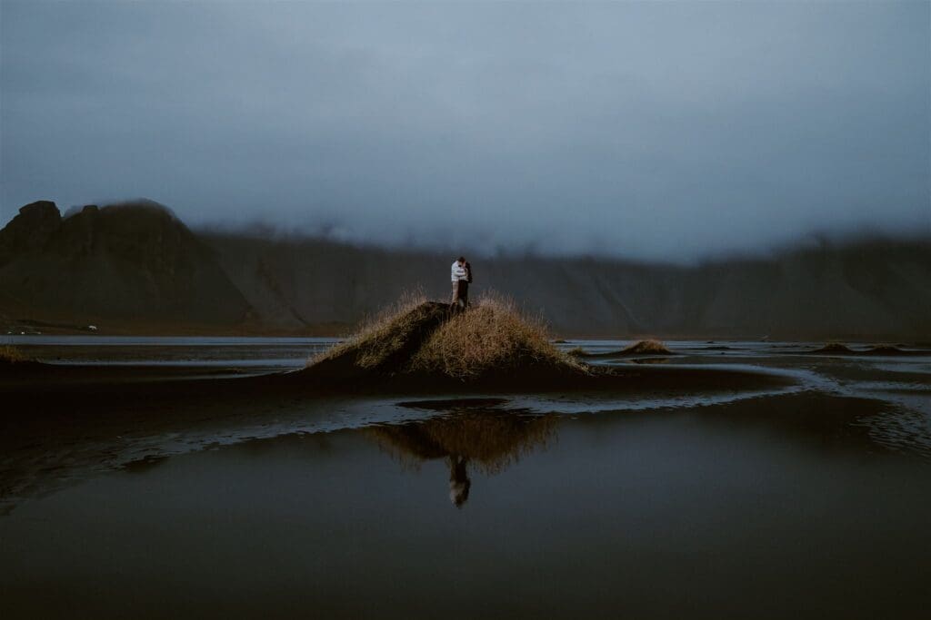 A couple stands on a black sand dune in Stokksnes, Iceland, with Vestrahorn mountain behind them covered in clouds