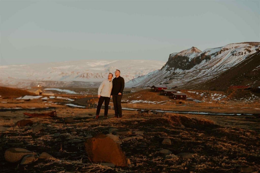 A gay couple looks at a raven flying overhead, as the soft winter light illuminates them and a winter backdrop of snow mountains and golden and black hues behind them
