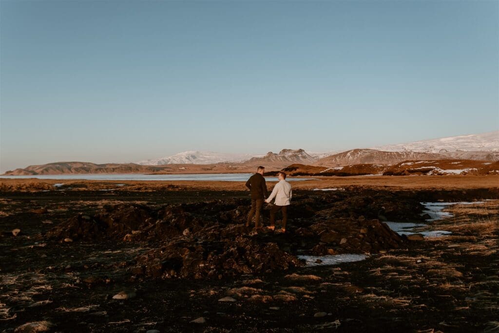 A couple walks hand in hand across Iceland's black sand beach in winter, with snowy mountains in the distance