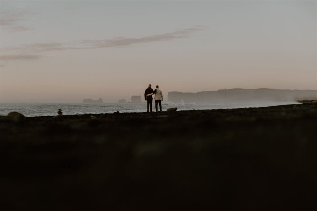 A couple standing on a black sand beach, arm in arm, overlooking Dyrholaey sea cliffs in the distance