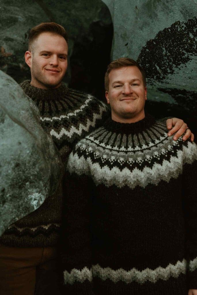 Gay couple wearing Icelandic lopapeysa, smiling together in an ice cave for their elopement