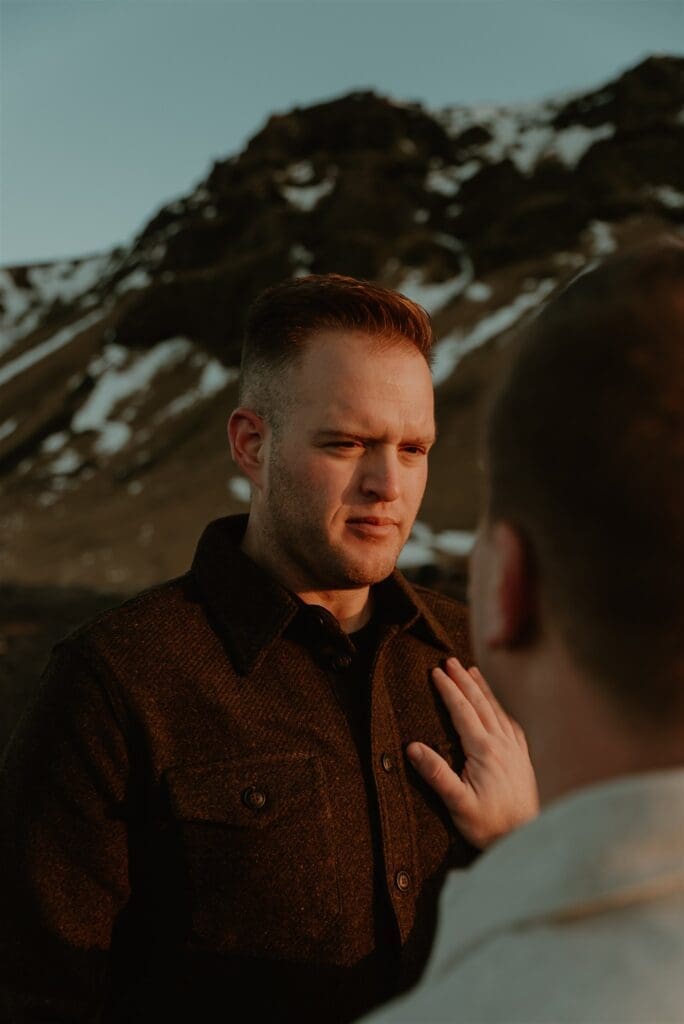 A man puts his hand over his fiancé's heart with soft golden sunrise light on his face, and a snow-capped mountain landscape behind him