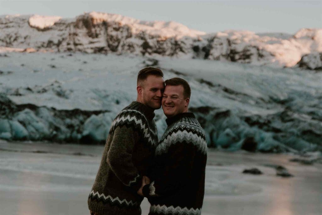 Male couple wearing lopapeysa embrace each other while looking at the camera and smiling