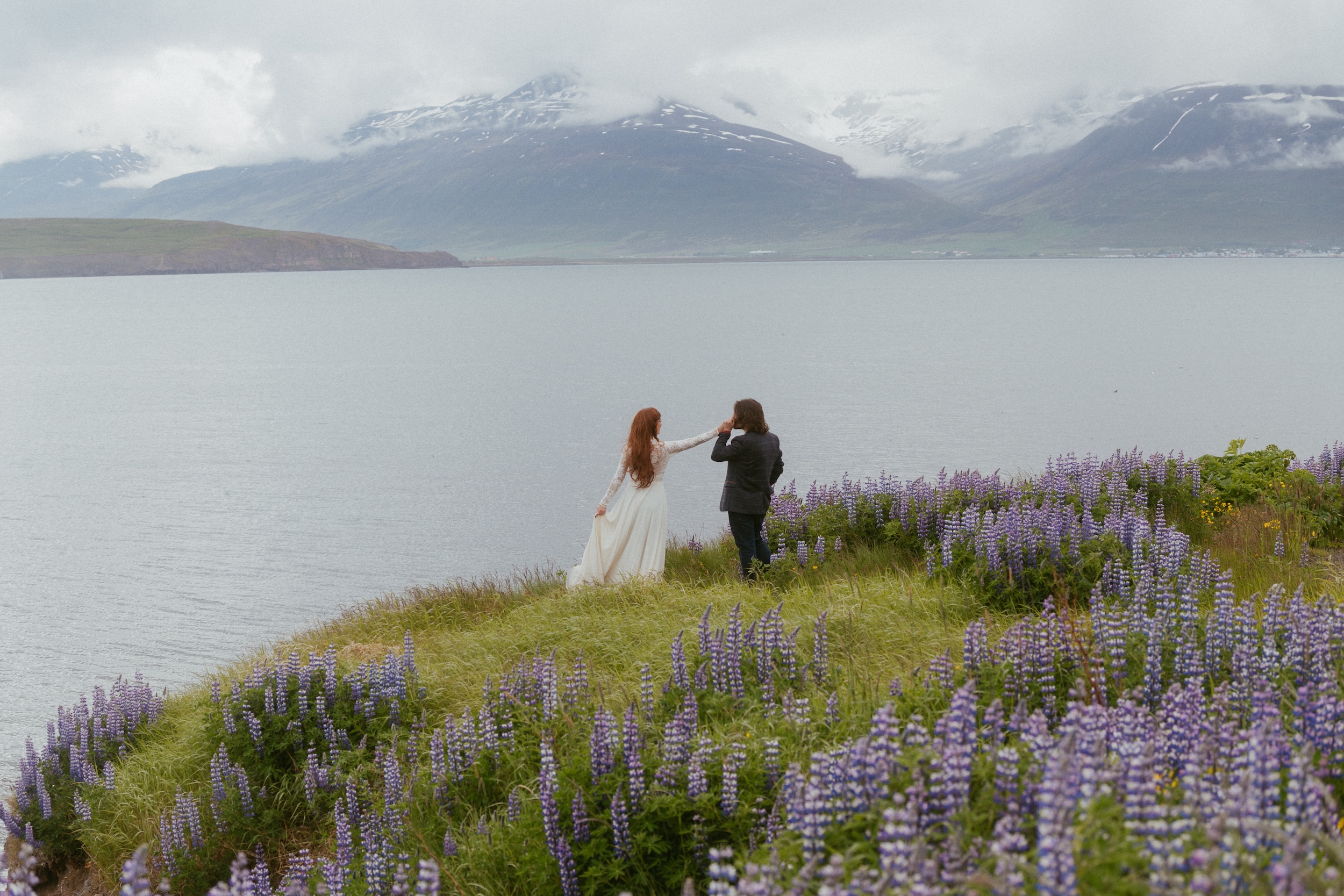 Iceland wedding couple standing on the edge of a pcturesque ocean cliff cilled with lupines and a mountain landscape in the background