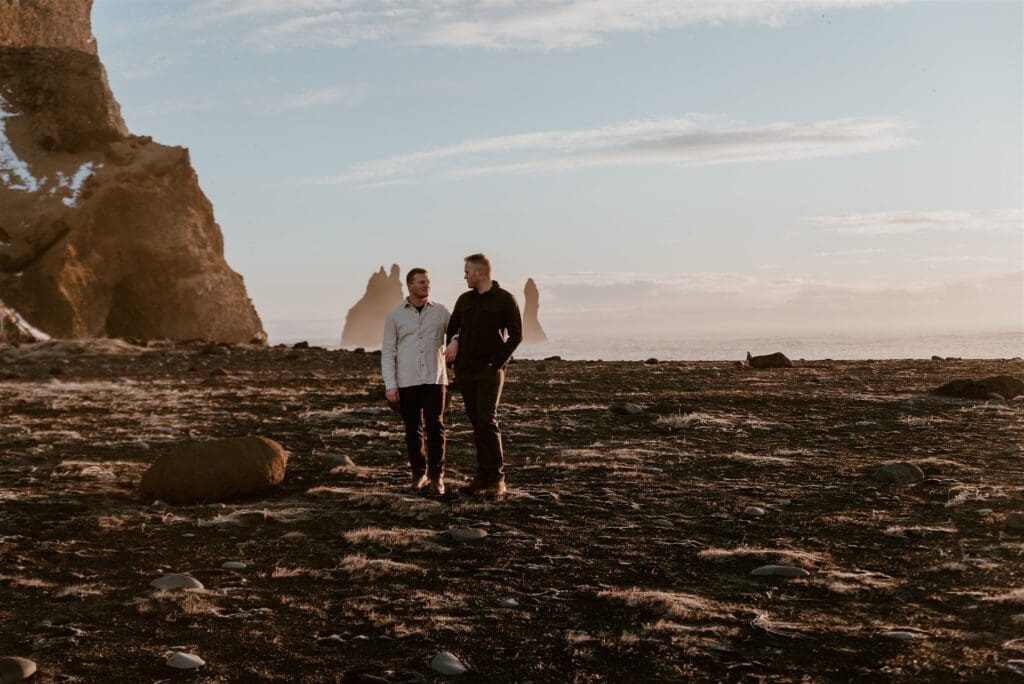 Gay couple walking arm in arm on a black sand beach in Iceland with giant sea stacks positioned behind them