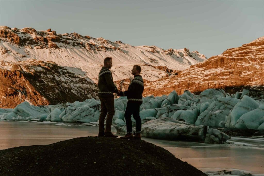 Male couple holds hands while standing on black lava sands underneath a glacier golden with light.