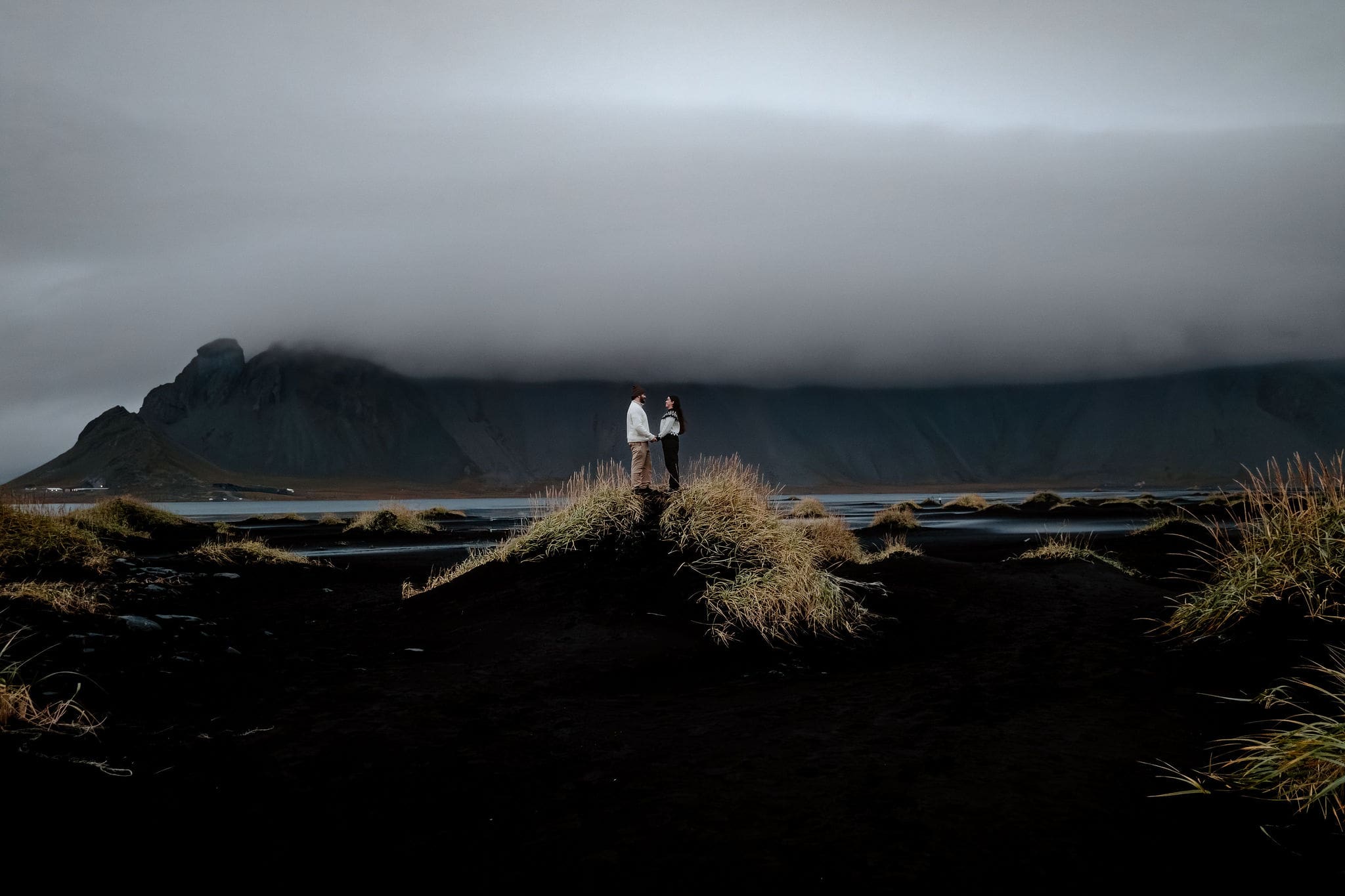 Engaged couple in Iceland atop a black sand beach dune at Stokksnes with a cloud-covered Vestrahorn behind them