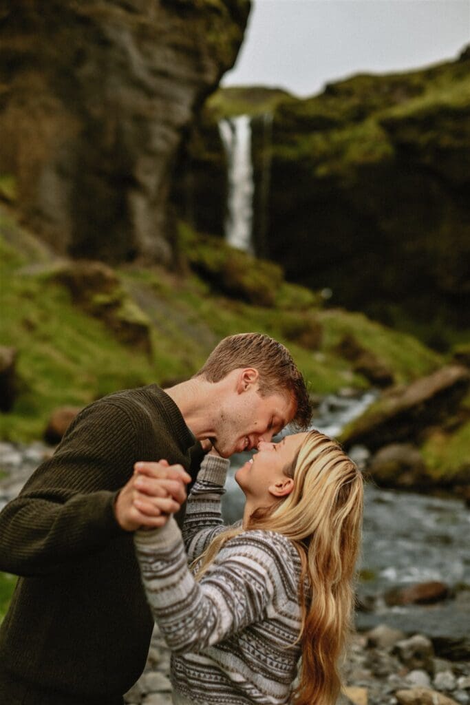 Couple kisses and holds hands after a surprise proposal with an Iceland waterfall behind them