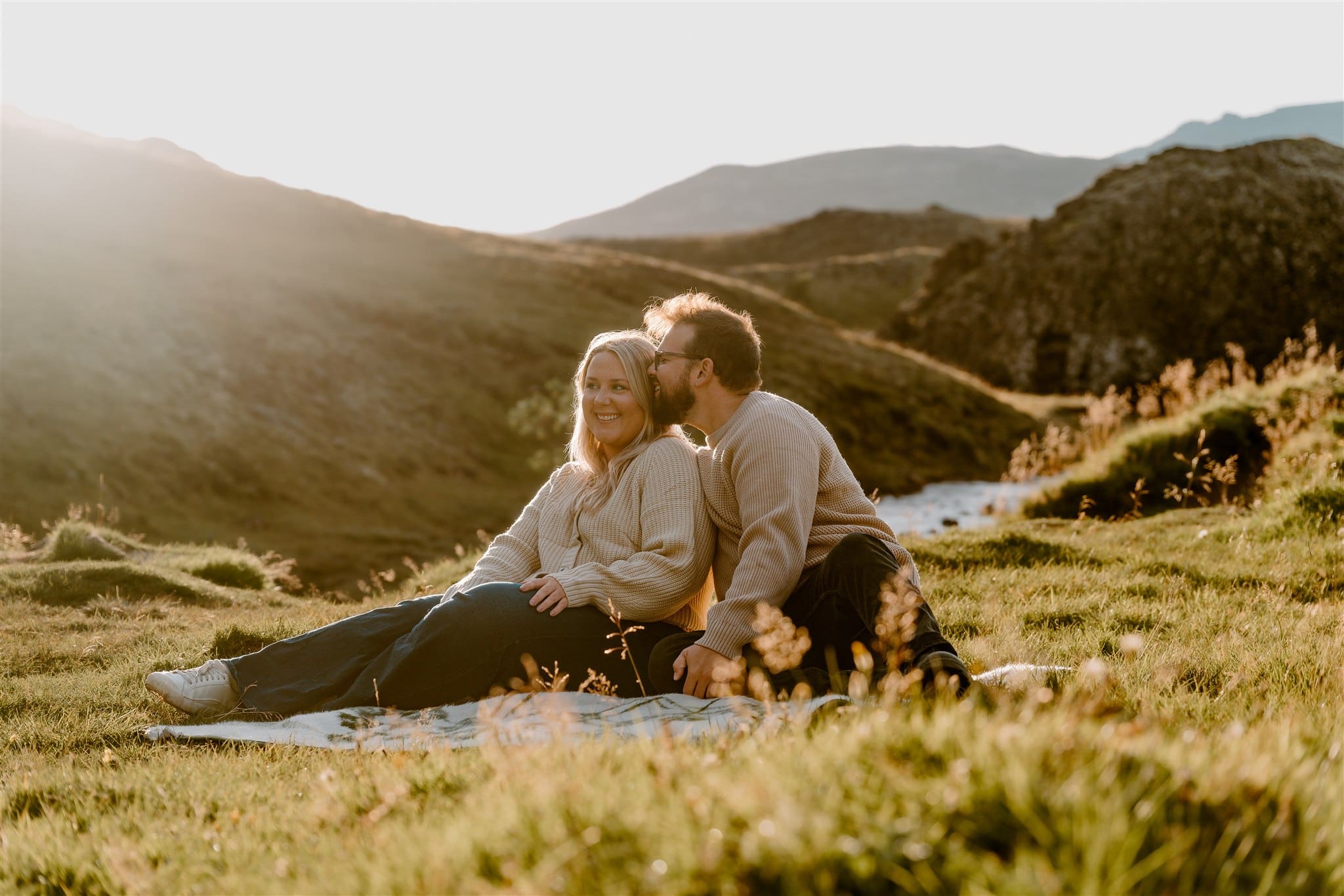 Happy, in-love couple sits in golden grass in a meadow with mountain landscape behind them