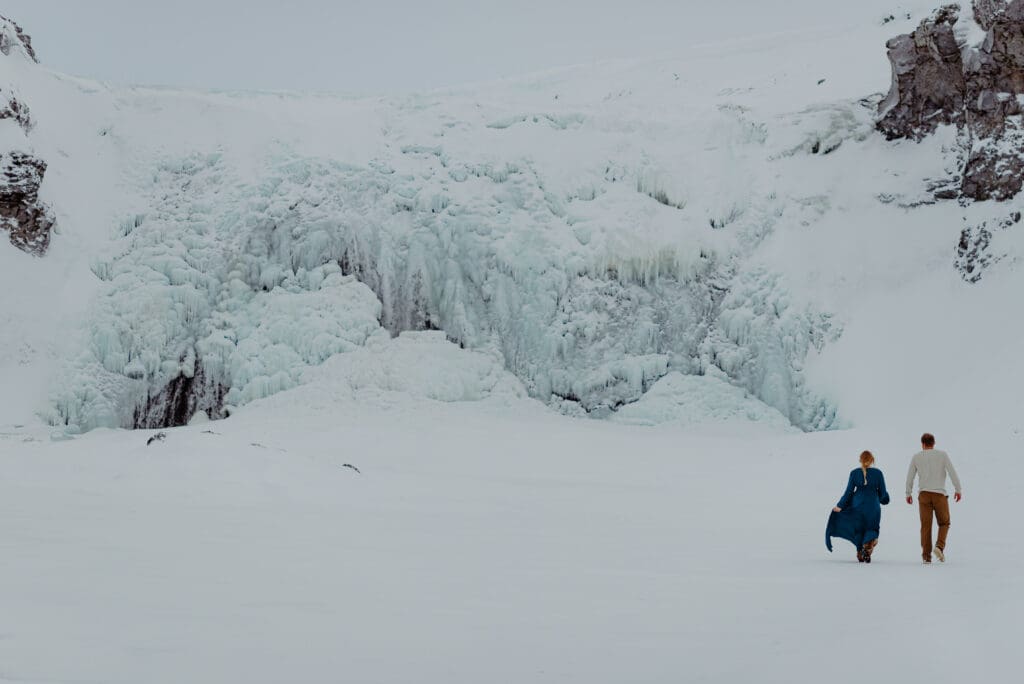 Couple walking across a frozen glacier landscape in Iceland beneath massive ice formations, cinematic winter adventure session near an Iceland glacier