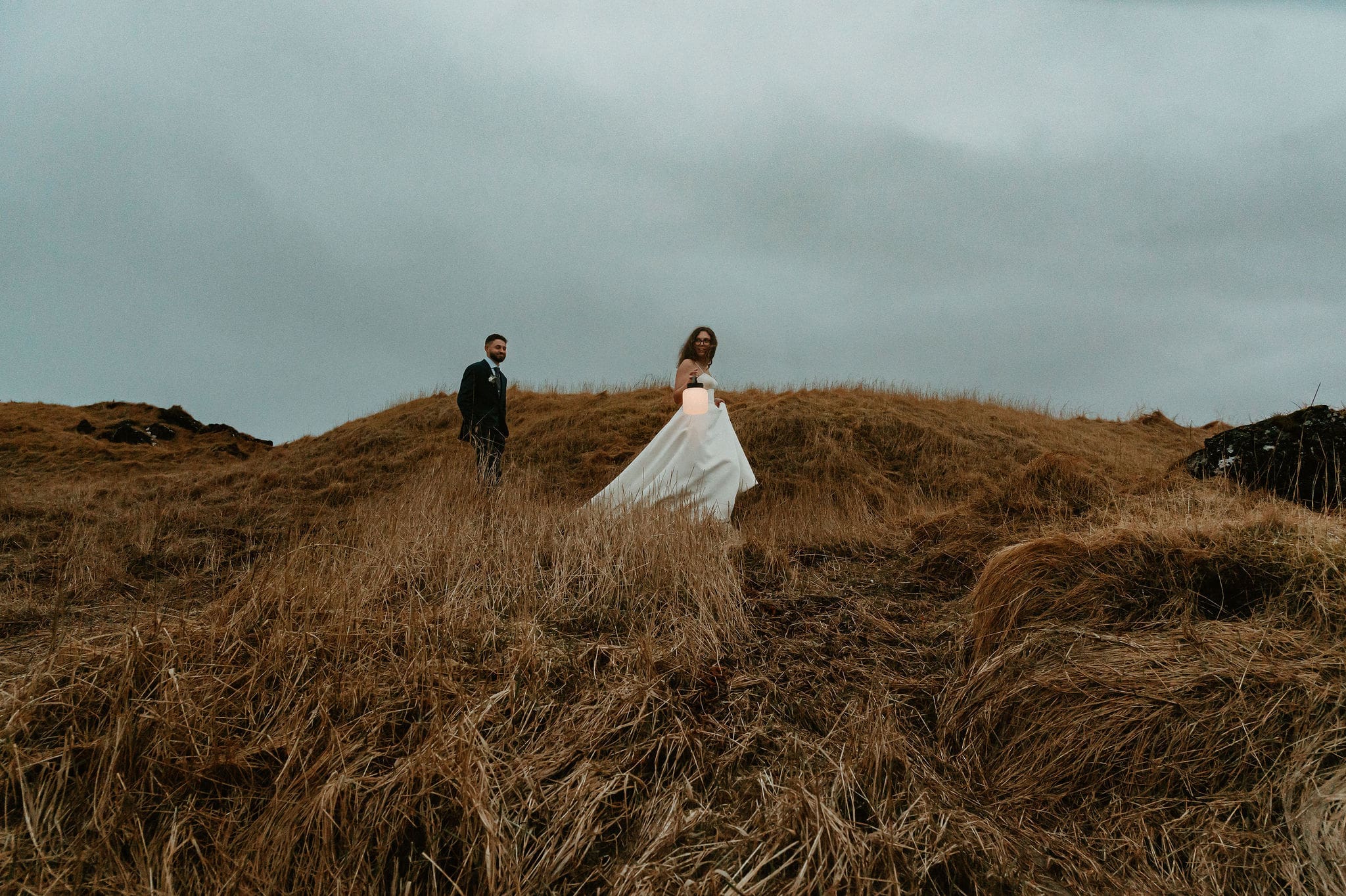 Bride and groom walking through windswept golden grass hills during an Iceland elopement, bride holding lantern in remote lava field landscape under moody overcast sky