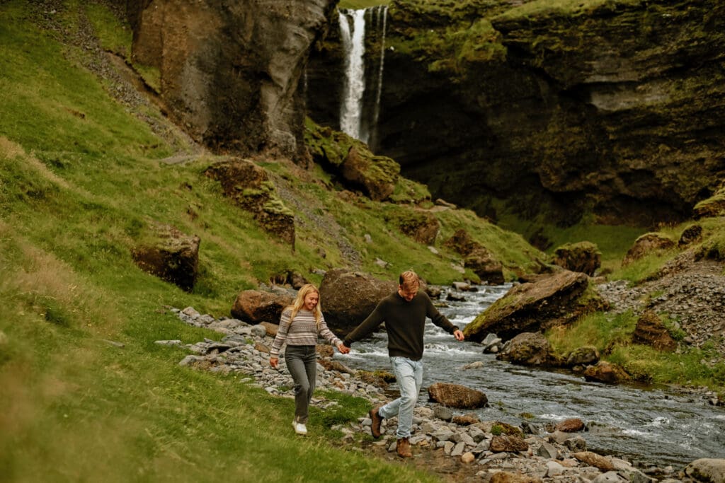 Couple holding hands and walking next to a creek that is fed by the waterfall Kvernufoss in Iceland