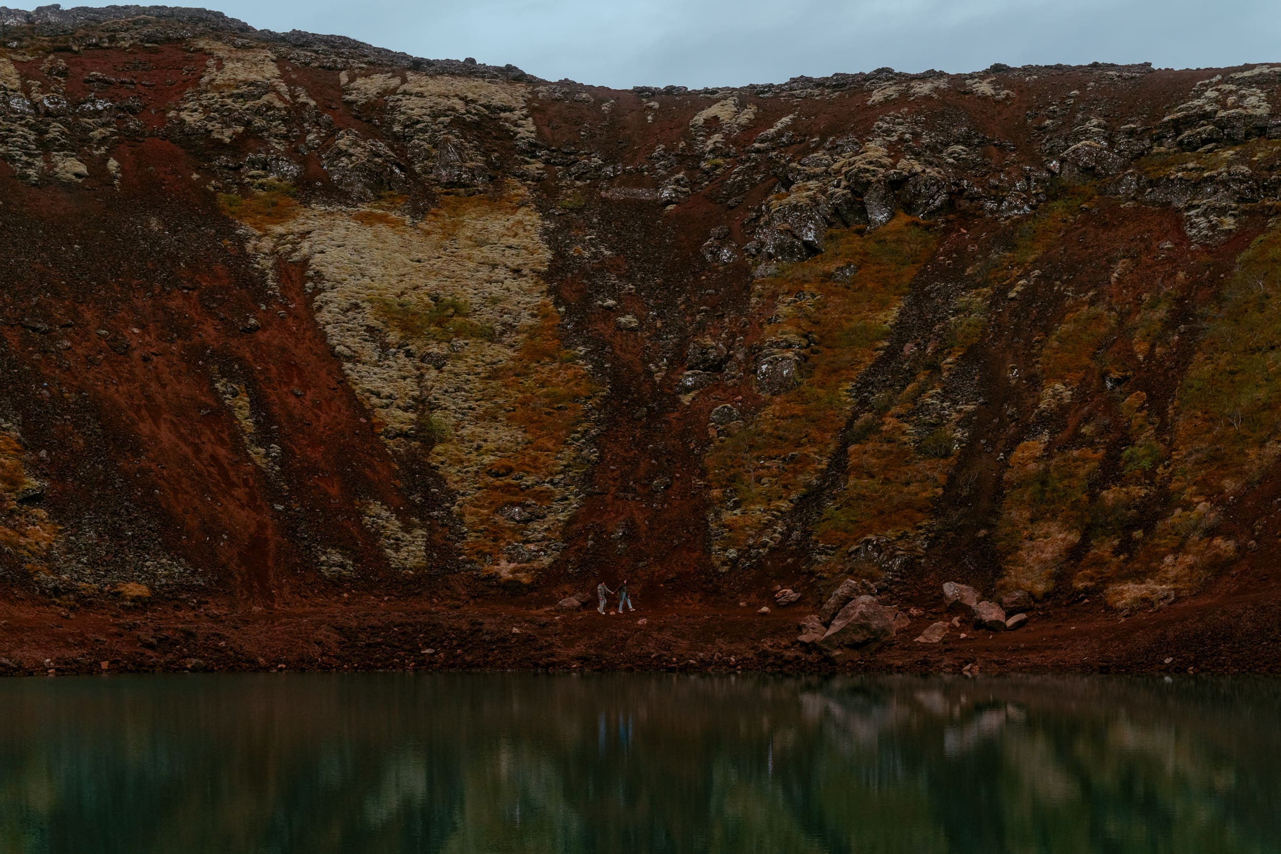 Otherworldly landscape image of elopement couple walking across a vast volcanic landscape inside a volcano crater, with red rock and green moss surrounding them and the turquoise crater lake in the foreground